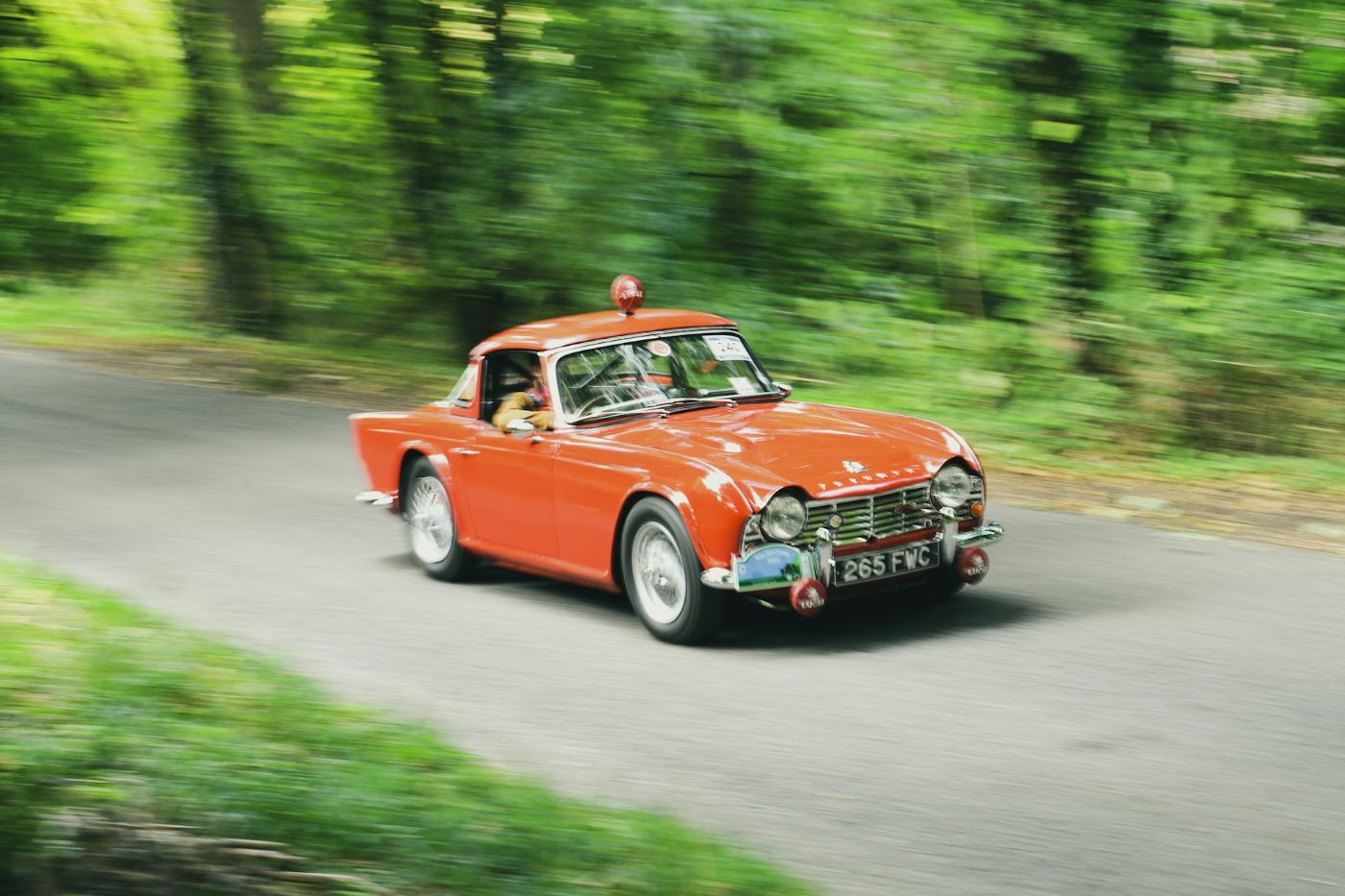 Triumph TR4 en mouvement sur route sinueuse avec arrière-plan flouté, illustrant le plaisir de conduite d'un roadster anglais classique.