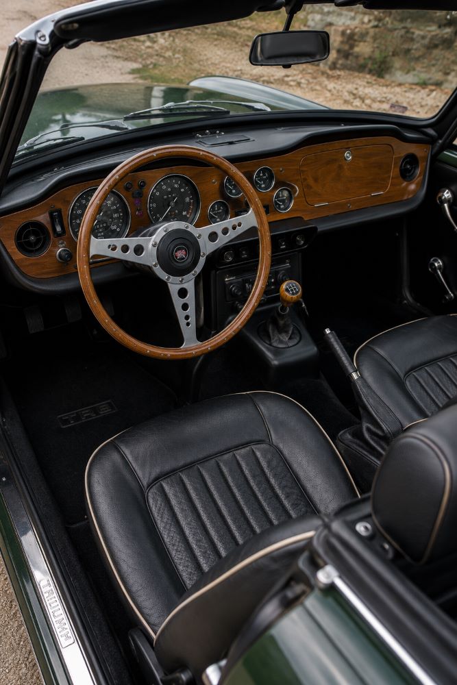 Habitacle d'une Triumph TR6 avec volant trois branches, tableau de bord en bois et compteurs Smiths, typique des roadsters britanniques des années 1970.