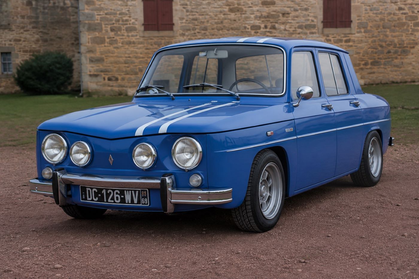 Renault 8 Gordini bleu de France vue trois-quarts avant avec bandes blanches, stationnée sur une route de campagne.