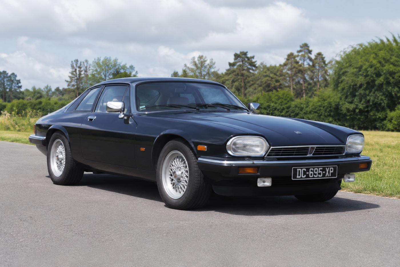 Jaguar XJS V12 bleu foncé photographiée de trois quarts avant sur une route de campagne. Cette vue met en valeur la silhouette élégante, le long capot et le design emblématique du grand coupé britannique.