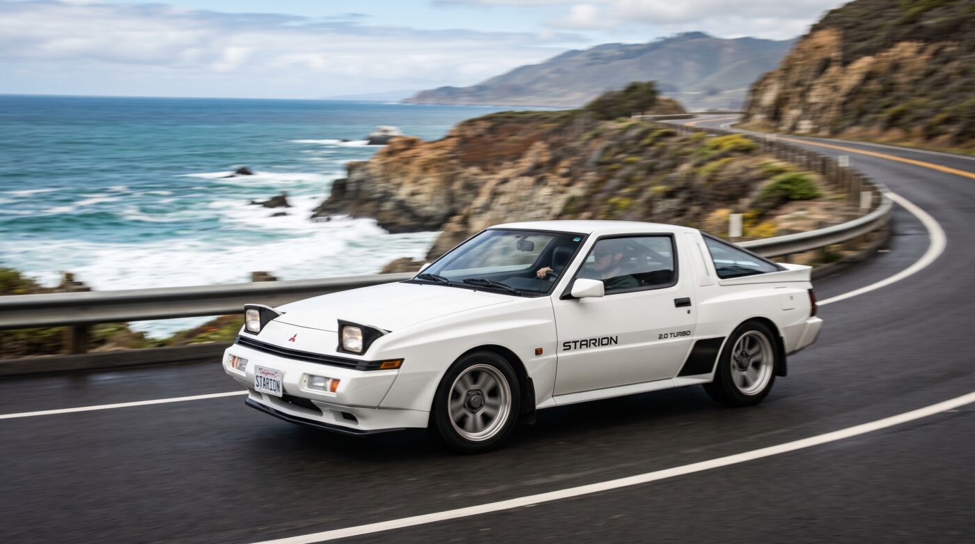 Mitsubishi Starion 1982 blanche en plein essai routier sur une route sinueuse, illustrant ses performances et son comportement dynamique.